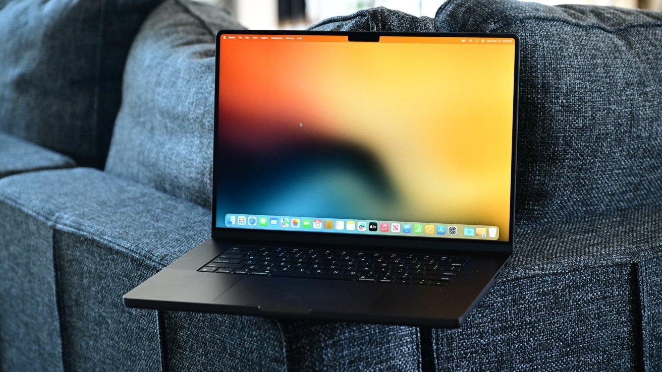 16palcový MacBook Pro Space Black od Apple s barevnou rozmazanou tapetou otevřený na šedé látkové pohovce.