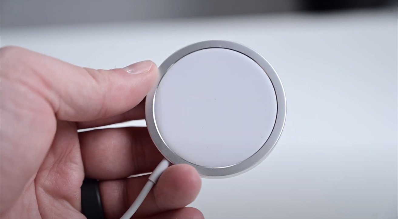 Ruka držící Apple MagSafe Charger s připojeným kabelem na rozmazaném světlém pozadí.