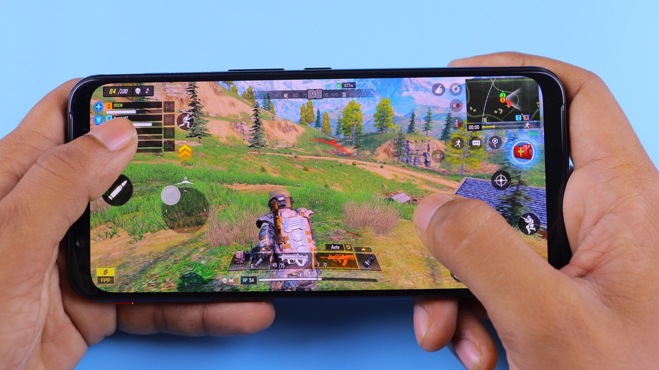 Ruce držící smartphone hrající hru Battle Royale s ovládacími prvky na obrazovce a viditelnou digitální krajinou.