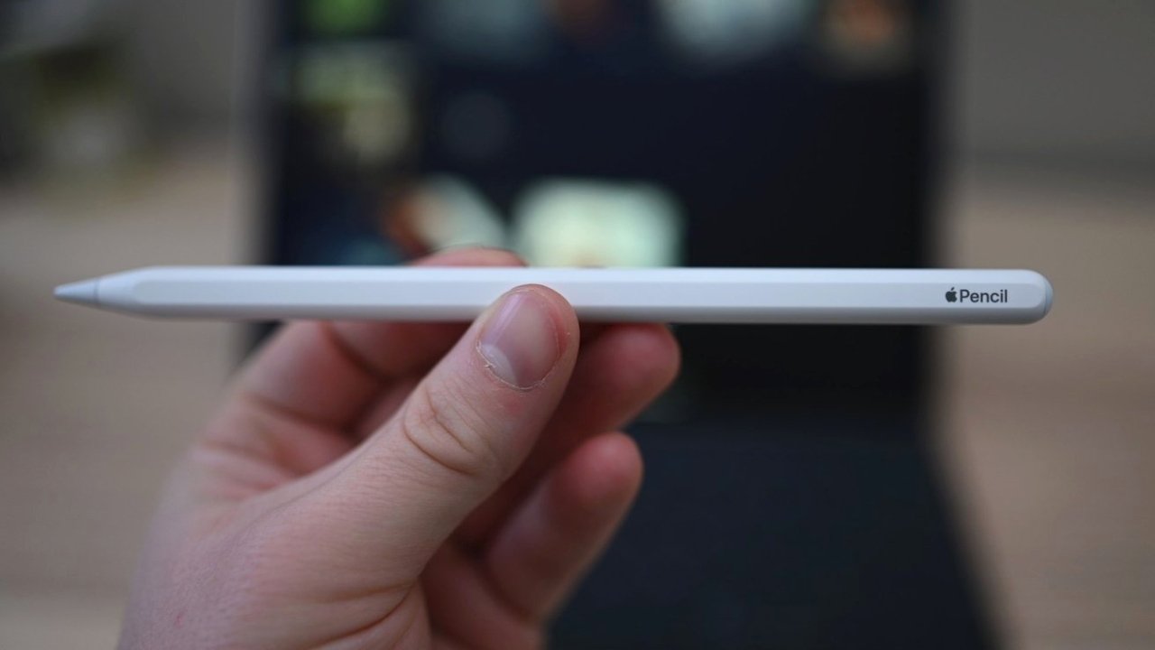 Detail ruky držící v centru pozornosti Apple Pencil s rozmazaným pozadím s tabletem.