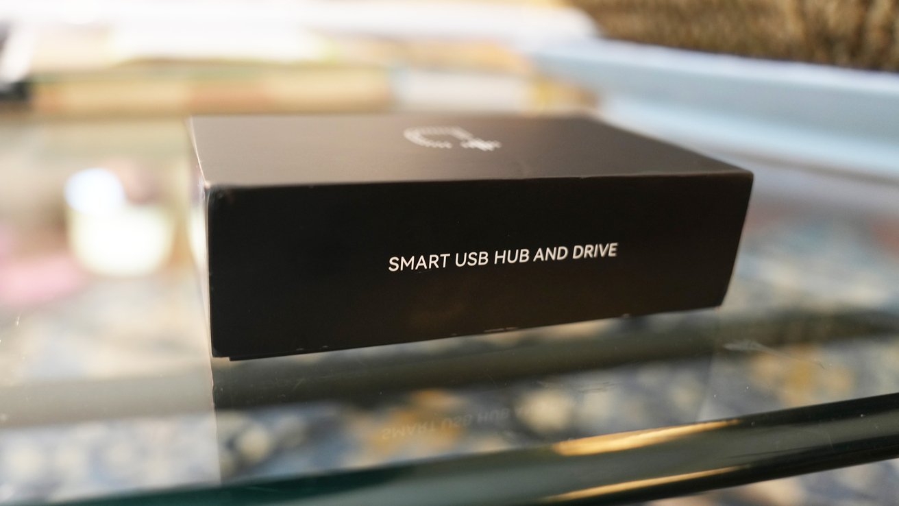 Balení dokovací skříně na skleněném stole označené bílým textem „SMART USB HUB AND DRIVE“.  Rozmazané pozadí.