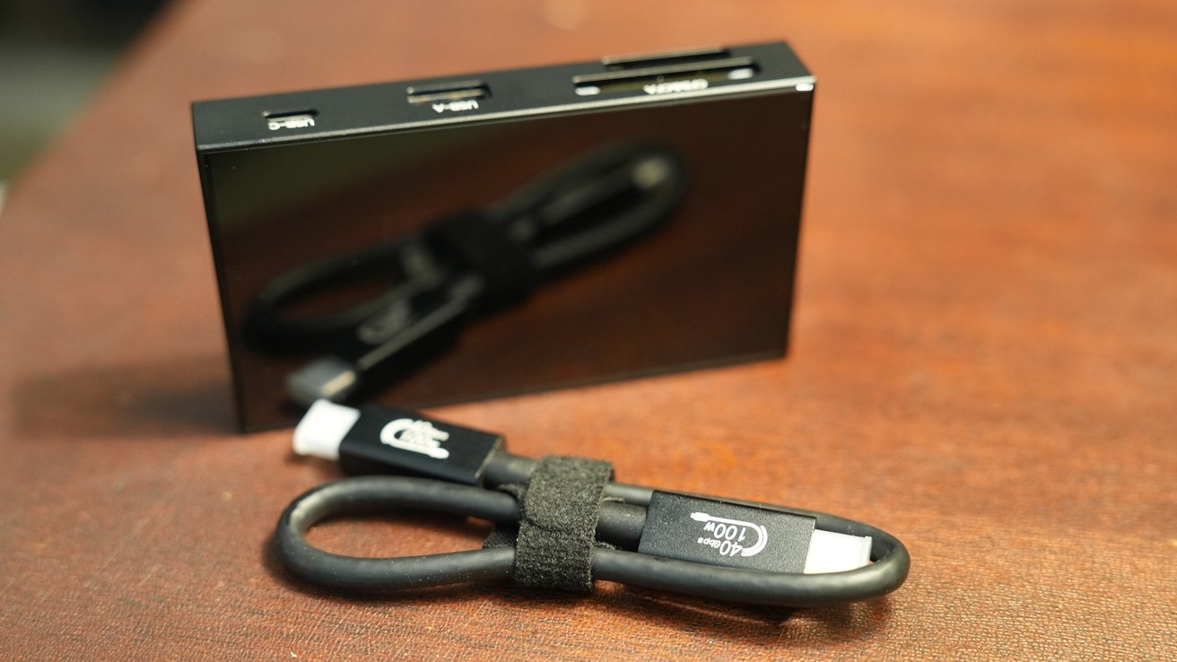 Černý multiportový adaptér se sloty pro USB-C, USB-A a SD karty na hnědém povrchu, vedle stočeného černého nabíjecího kabelu.