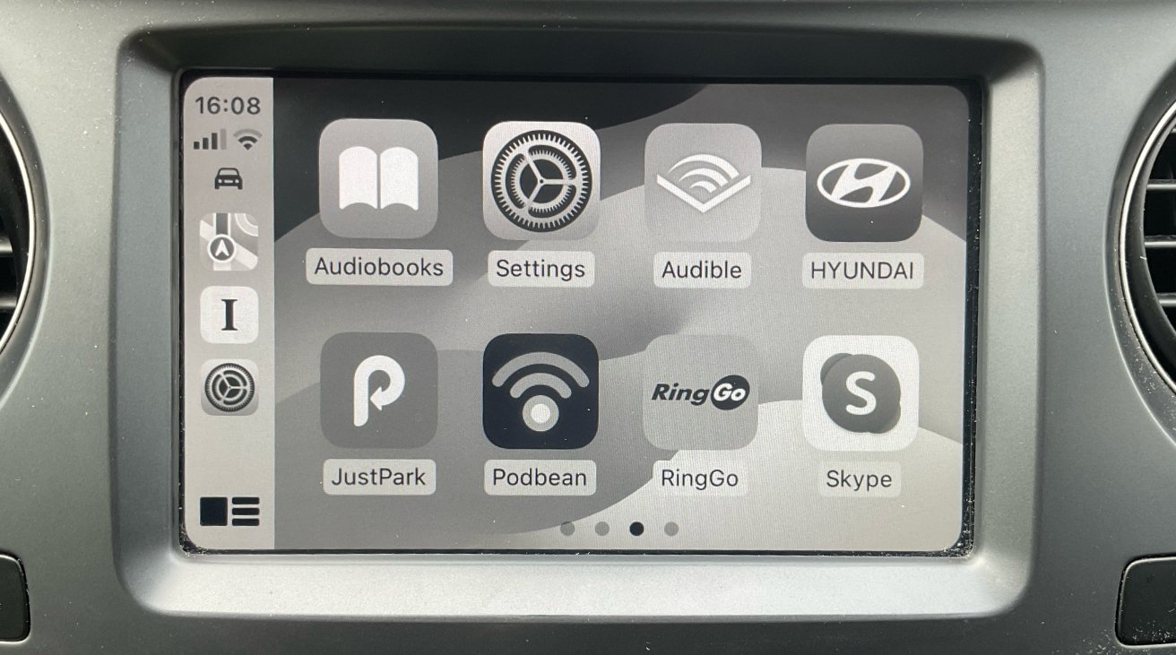 Displej automobilového informačního a zábavního systému zobrazující ikony aplikací včetně Audiobooks, Settings, Audible, Hyundai, JustPark, Podbean, RingGo a Skype.