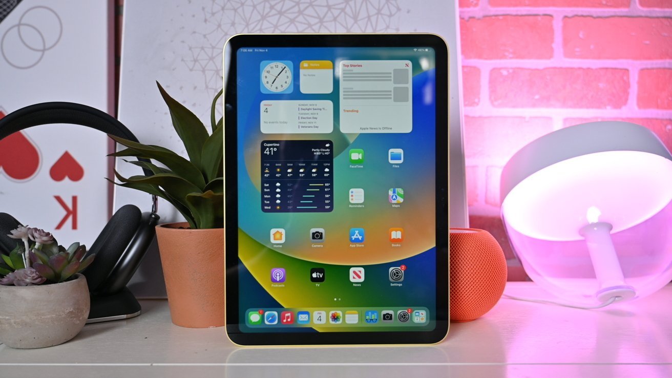 iPad 10th Generatin na stojanu zobrazující aplikace na domovské obrazovce, lemovaný rostlinami a technickými doplňky na stole.