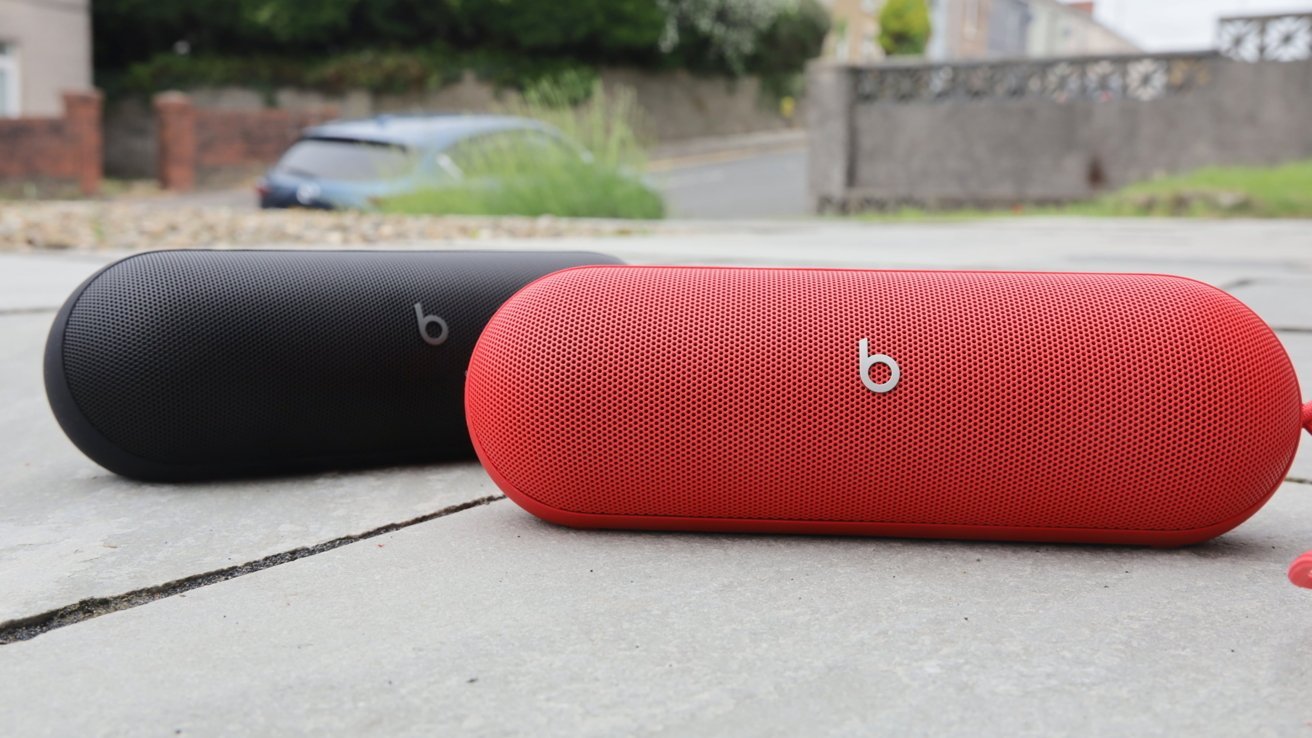 Dva přenosné reproduktory Beats Pill, jeden černý a jeden červený, umístěné na venkovním chodníku s rozostřeným pozadím ulice.