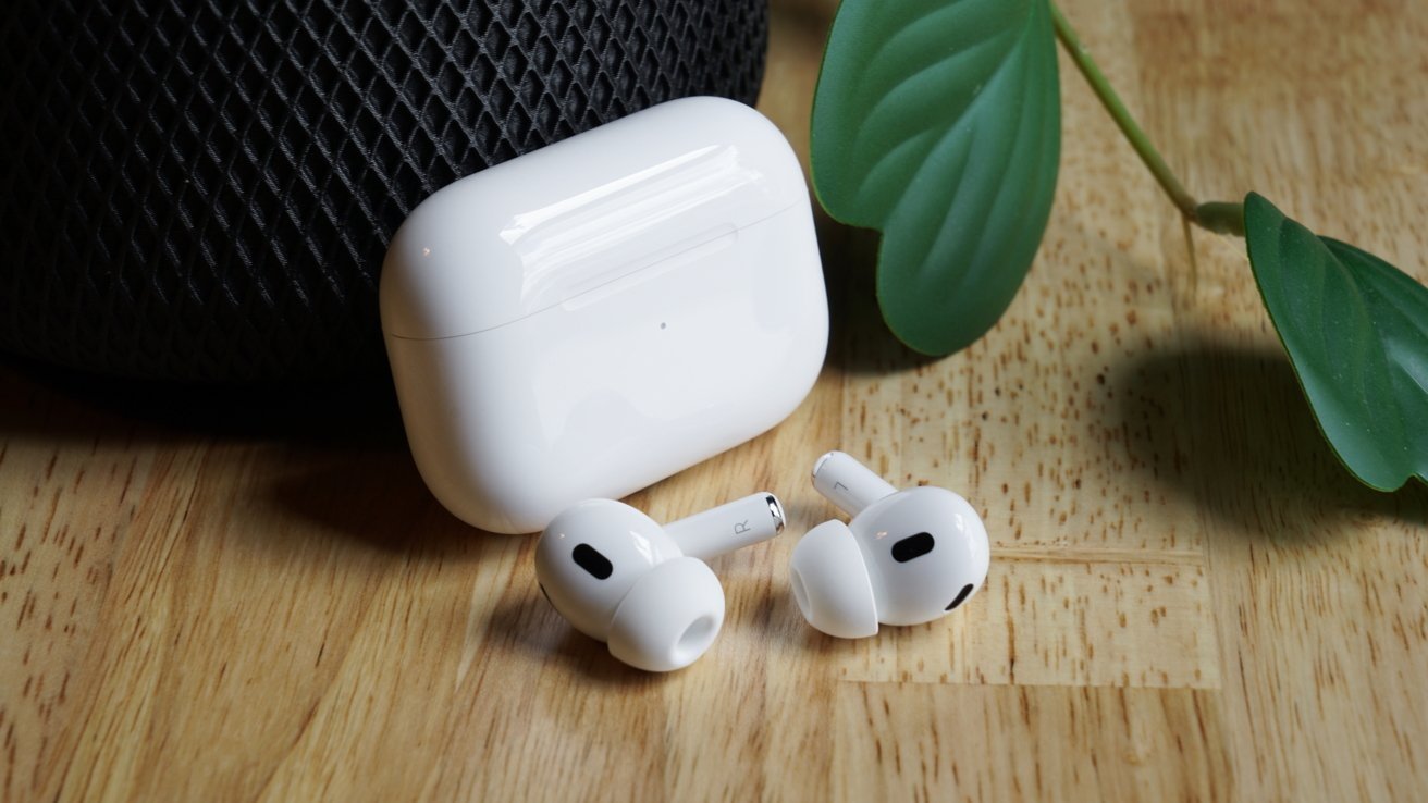 AirPods vedle jejich otevřeného nabíjecího pouzdra na dřevěném povrchu s rostlinným listem a reproduktorem HomePod v pozadí.