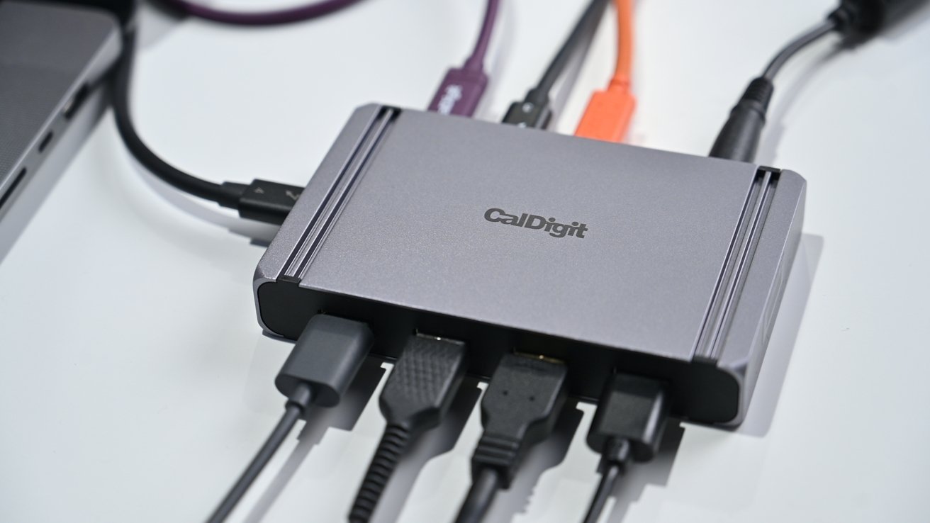 Kompaktní rozbočovač CalDigit s více připojenými kabely, včetně USB a HDMI, ležící vedle notebooku na bílém povrchu.