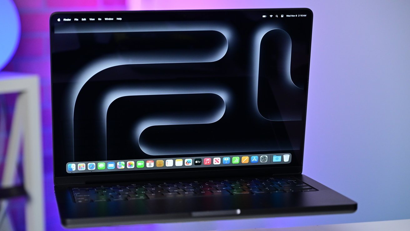 Laptop MacBook Pro s barevnou abstraktní tapetou, zářícími ikonami aplikací a tmavou klávesnicí na moderním stole.