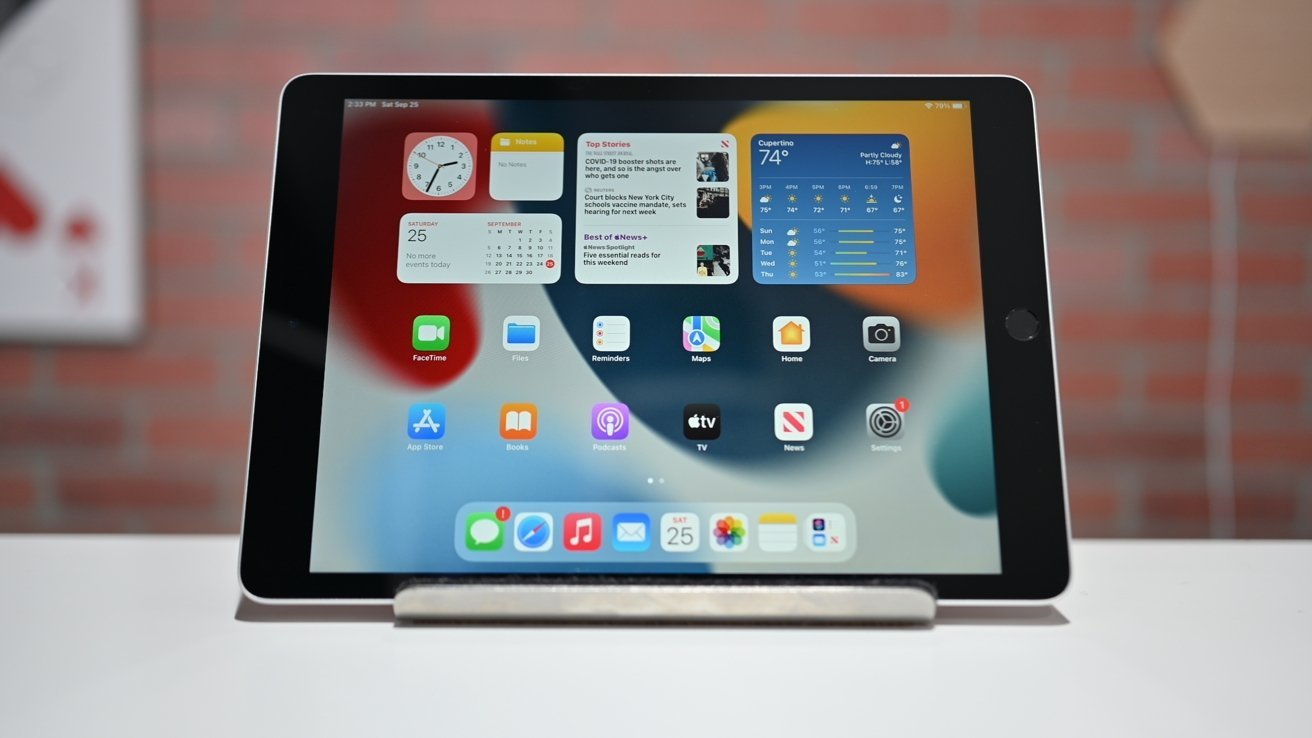 iPad 9. generace zobrazující různé aplikace a widgety na své domovské obrazovce, umístěný na stojanu proti rozmazanému pozadí cihlové zdi.