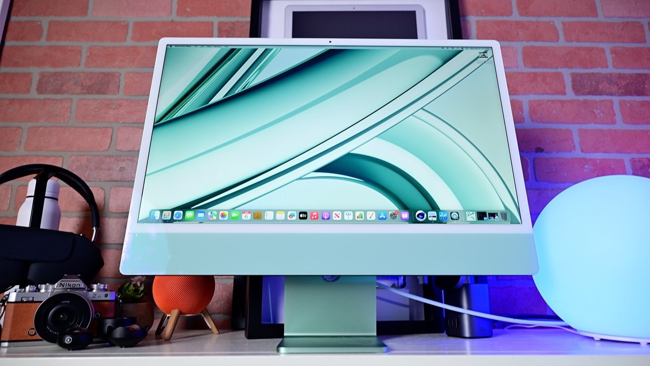 Počítač Apple iMac s pastelově zelenou obrazovkou na stole, obklopený fotoaparátem Nikon, sluchátky AirPods Max, lahví na vodu, oranžovým reproduktorem a zářící modrou koulí. Pozadí cihlové zdi.