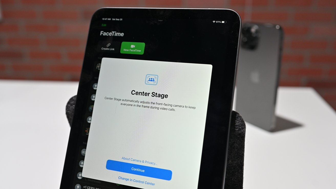 obrazovka iPadu zobrazující nastavení FaceTime Center Stage, optimalizující přední kameru pro videohovory; pozadí odhaluje rozmazaný iPhone na stojanu.