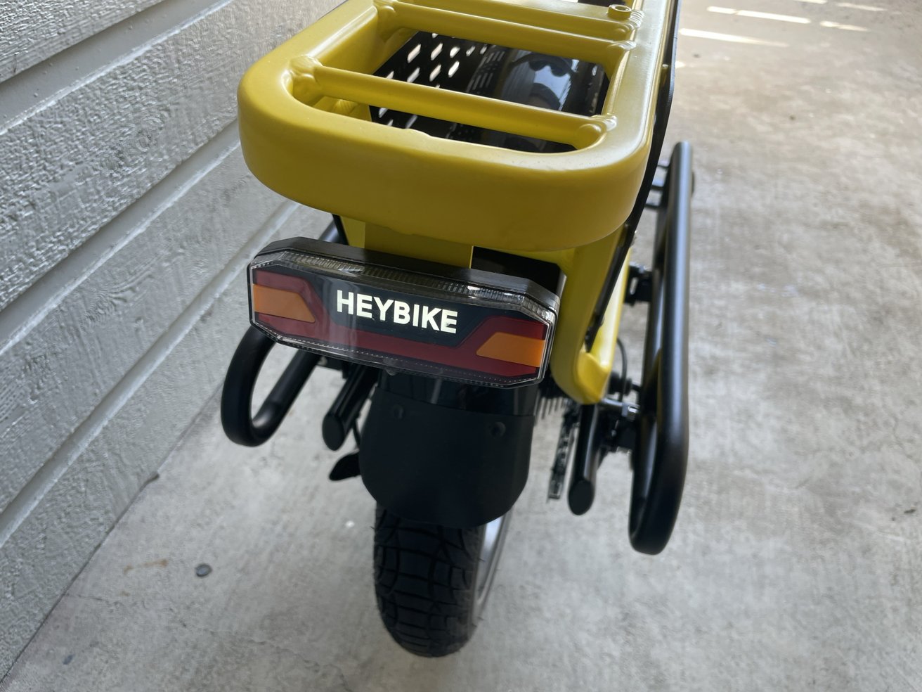 Pohled na žlutý Heybike zezadu, zobrazující zadní pneumatiku, blatník a nákladový nosič vedle texturované bílé stěny.