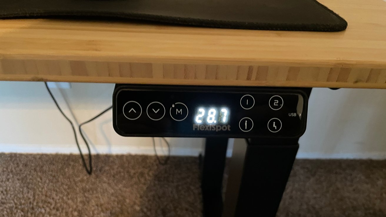 Nastavitelný stolní ovládací panel zobrazující 28,7 s tlačítky pro nastavení výšky a předvolby pod dřevěným stolem. V pozadí jsou vidět elektrické šňůry.
