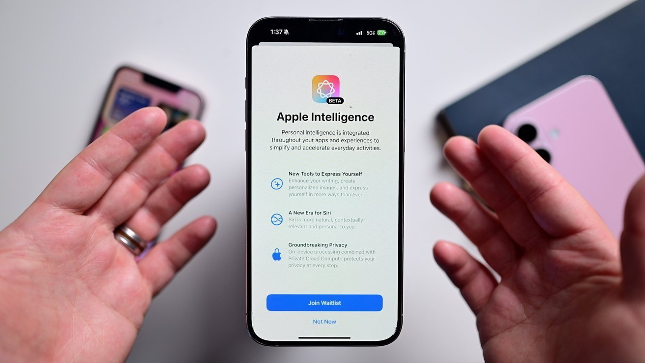 Ruce držící smartphone zobrazující obrazovku o Apple Intelligence beta, zdůrazňující nové nástroje, vylepšení Siri a soukromí. Na stole v pozadí jsou dva telefony.