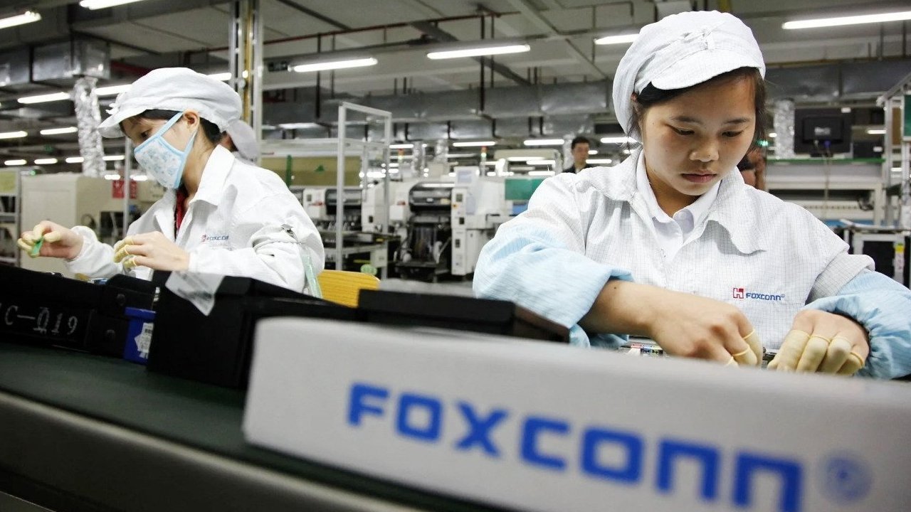 Tovární dělníci v bílých uniformách a čepicích montují elektroniku na výrobní lince, přičemž na zařízení je vidět slovo Foxconn.