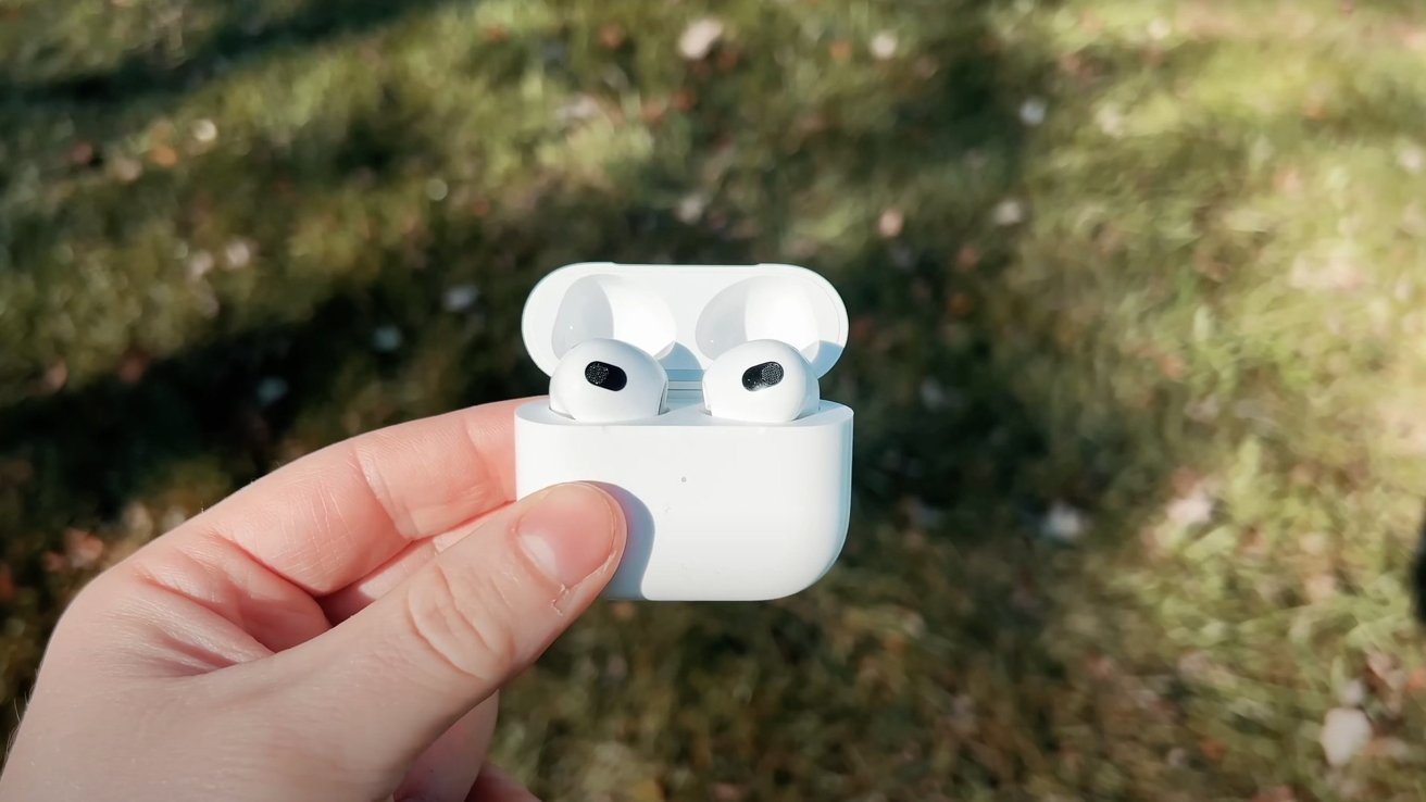 Ruka drží otevřené bílé nabíjecí pouzdro AirPods Pro 2 na travnatém pozadí.