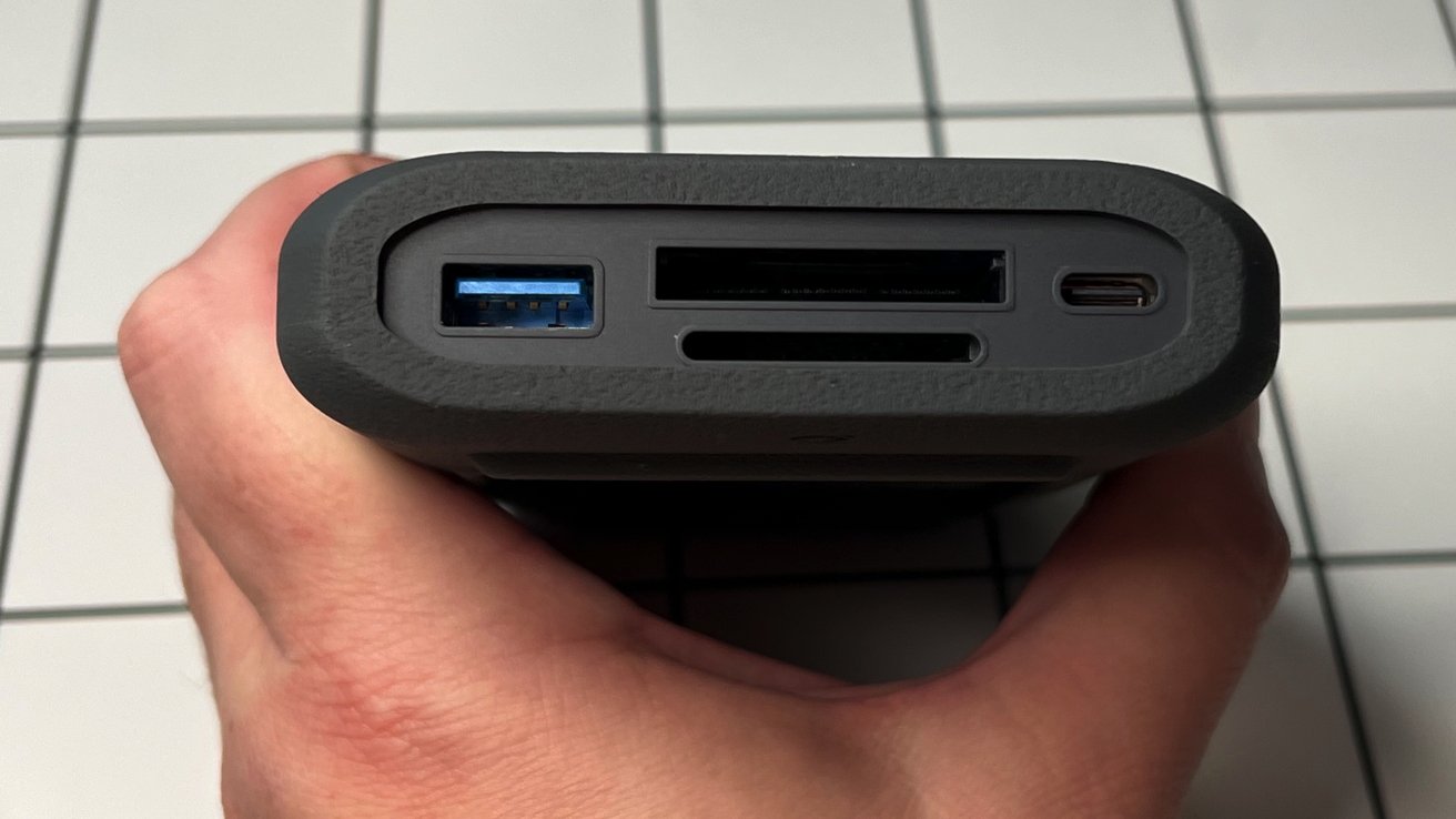 Ruka držící černé zařízení s porty včetně USB, SD karty a USB-C na dlaždicovém pozadí.