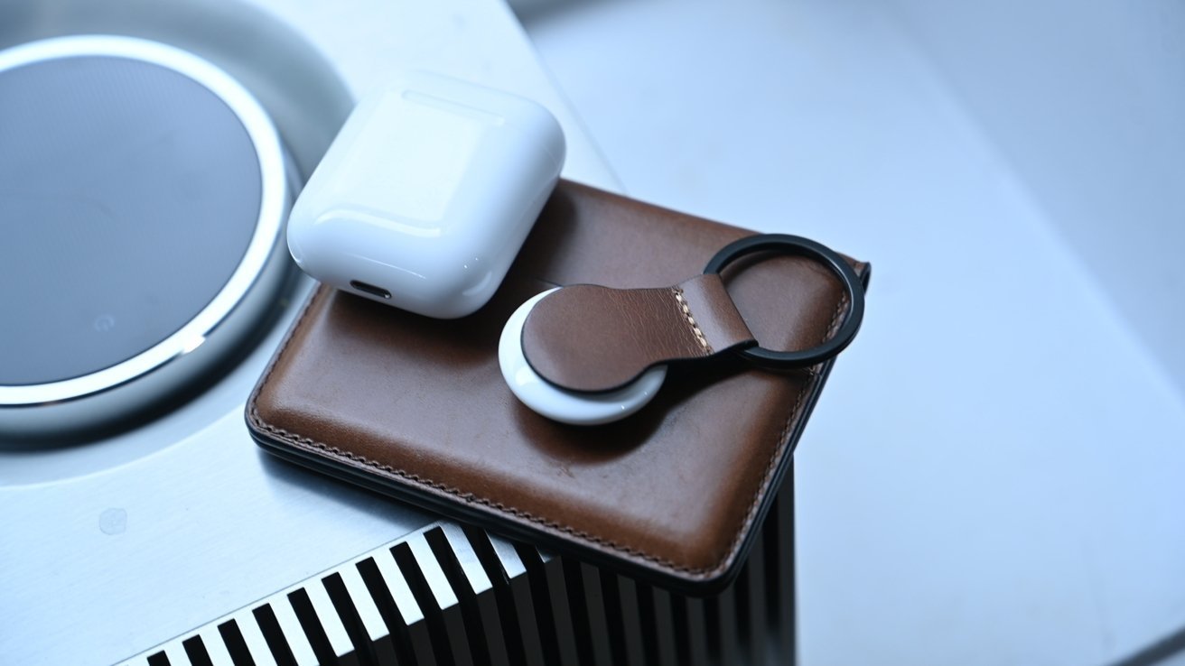 Hnědá kožená peněženka s pouzdrem AirPods a Nomad AirTag Leather Loop na stříbrném povrchu.