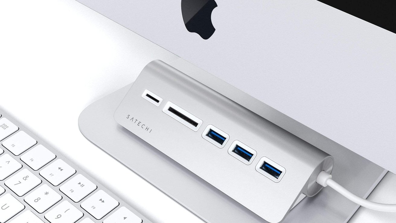 Stříbrný stolní USB hub s více porty pod monitorem, vedle bílé klávesnice.