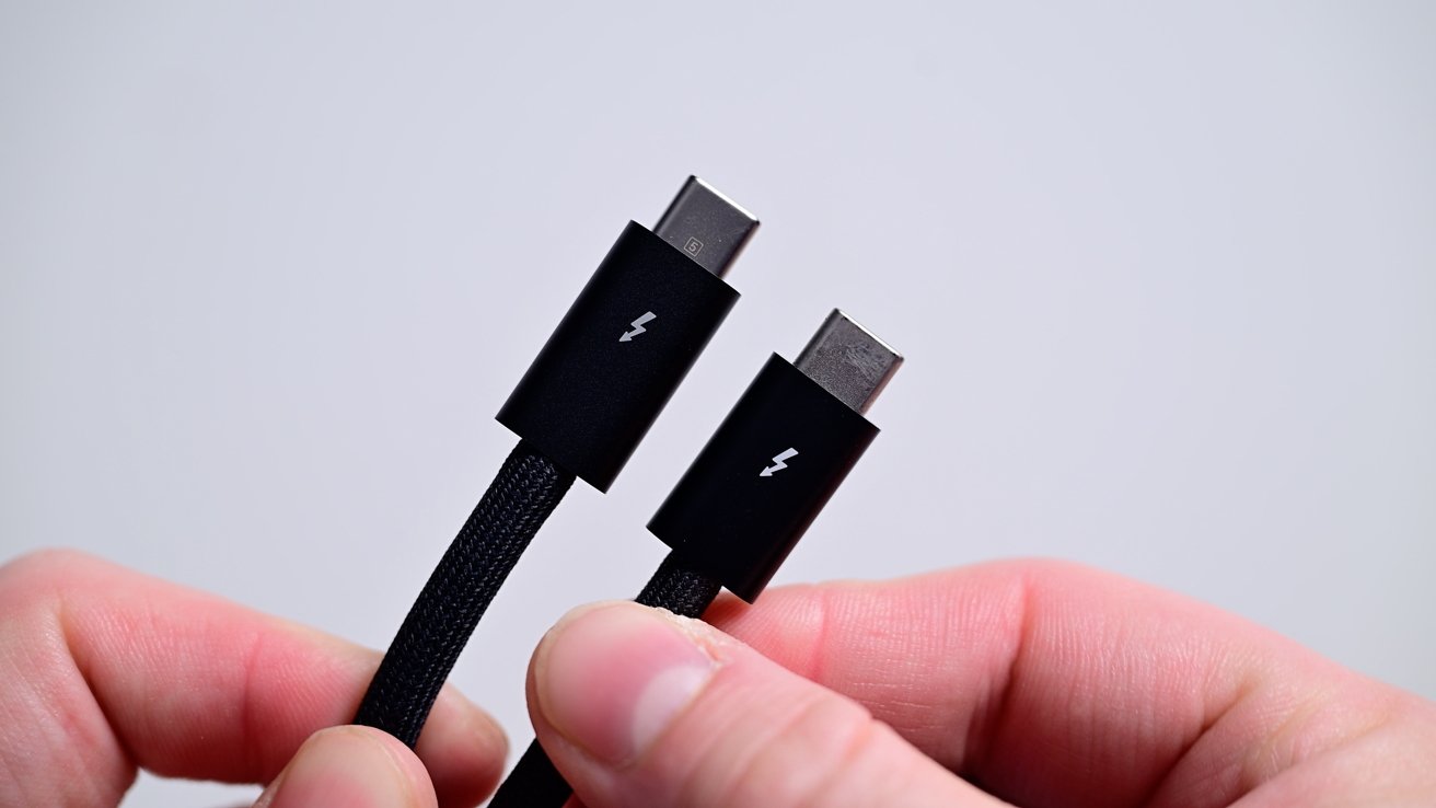 Dva černé kabely USB-C se symboly blesků držených mezi prsty na prostém pozadí.